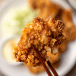 Morceau de karaage croustillant tenu par des baguettes japonaises, en gros plan, avec salade de chou et sauce en arrière-plan