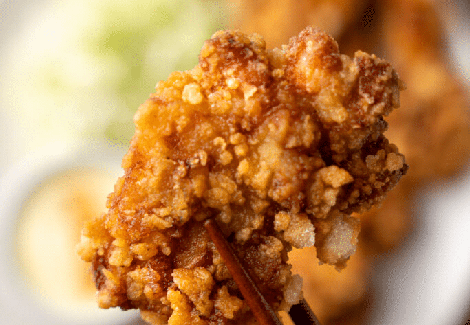 Morceau de karaage croustillant tenu par des baguettes japonaises, en gros plan, avec salade de chou et sauce en arrière-plan