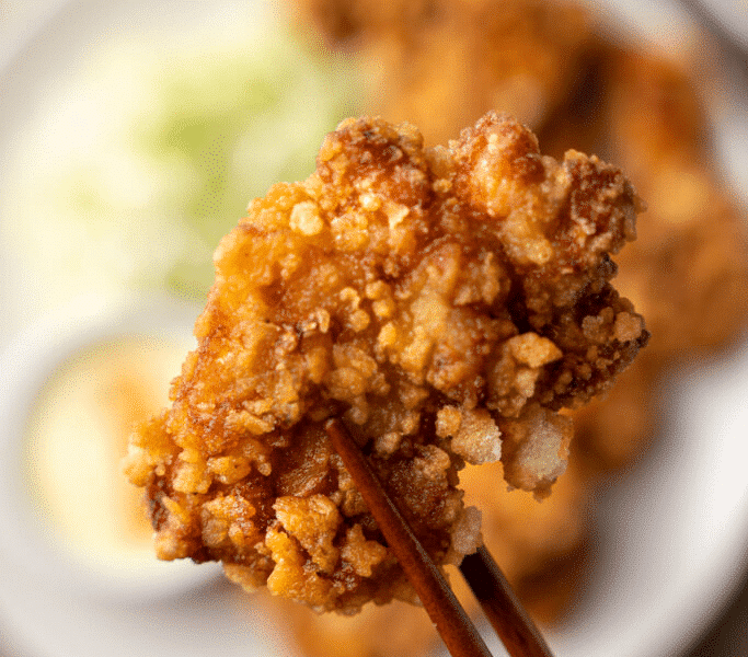 Morceau de karaage croustillant tenu par des baguettes japonaises, en gros plan, avec salade de chou et sauce en arrière-plan